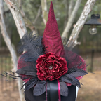 A gothic-style witch hat decoration featuring a tall pointed cone in deep burgundy velvet, a large matching burgundy flower, black feathers, and black mesh accents, attached to a black hat band with a ribbon.