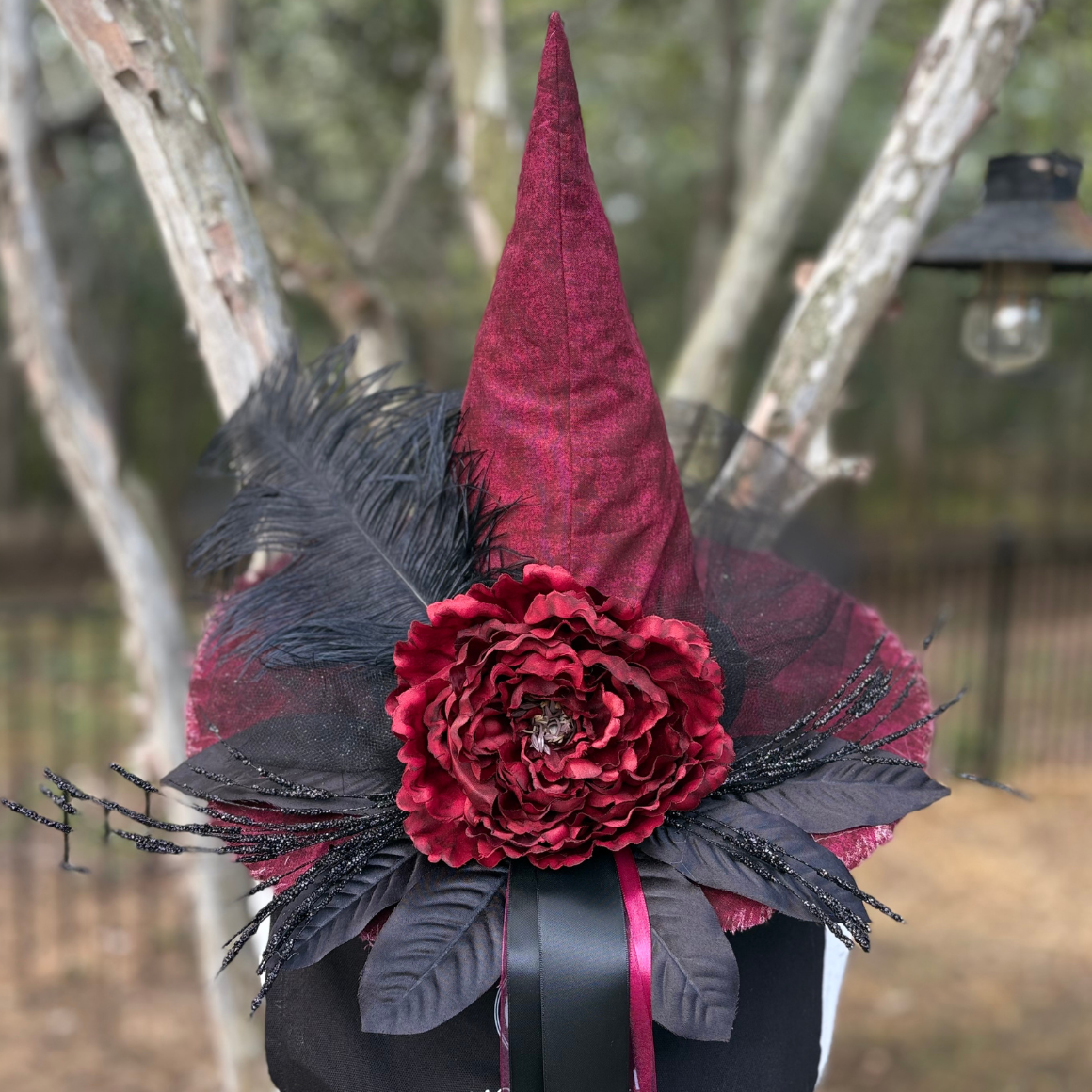 A gothic-style witch hat decoration featuring a tall pointed cone in deep burgundy velvet, a large matching burgundy flower, black feathers, and black mesh accents, attached to a black hat band with a ribbon.