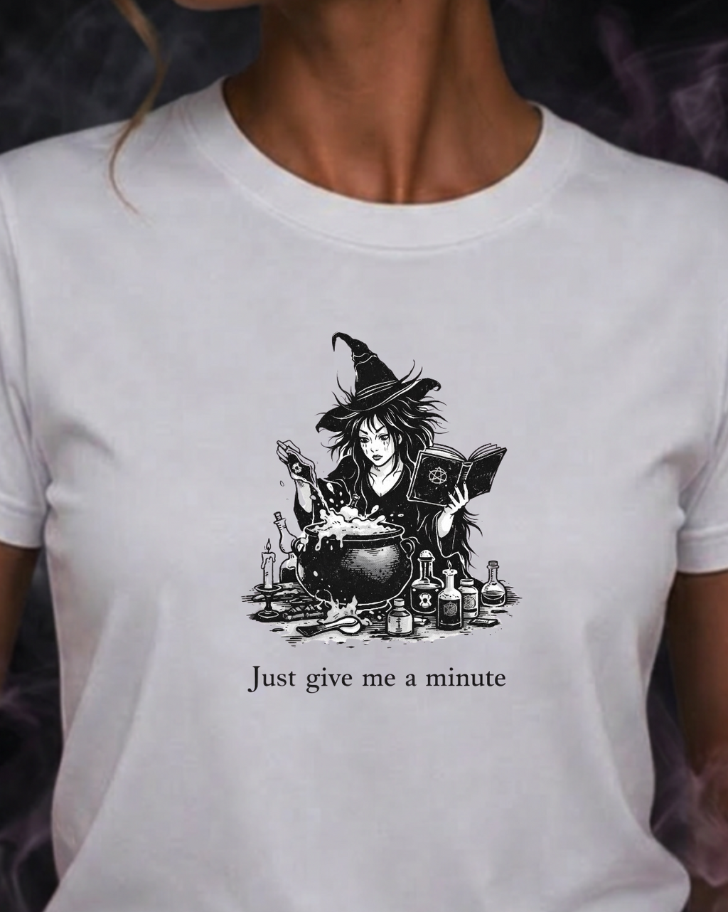 A woman wearing a white short-sleeve t-shirt with a black graphic of a witch and the text 'Just give me a minute' on the front, set against a dark smoky background.