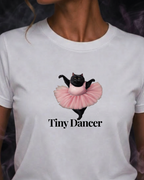 A woman wearing a white short-sleeve t-shirt with a graphic of a black cat in a pink tutu and the text 'Tiny Dancer' printed below it.