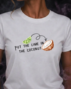 A woman wearing a white short-sleeve graphic t-shirt with the text 'PUT THE LIME IN THE COCONUT' and illustrations of a lime slice and a coconut. The background is dark with purple smoke effects.