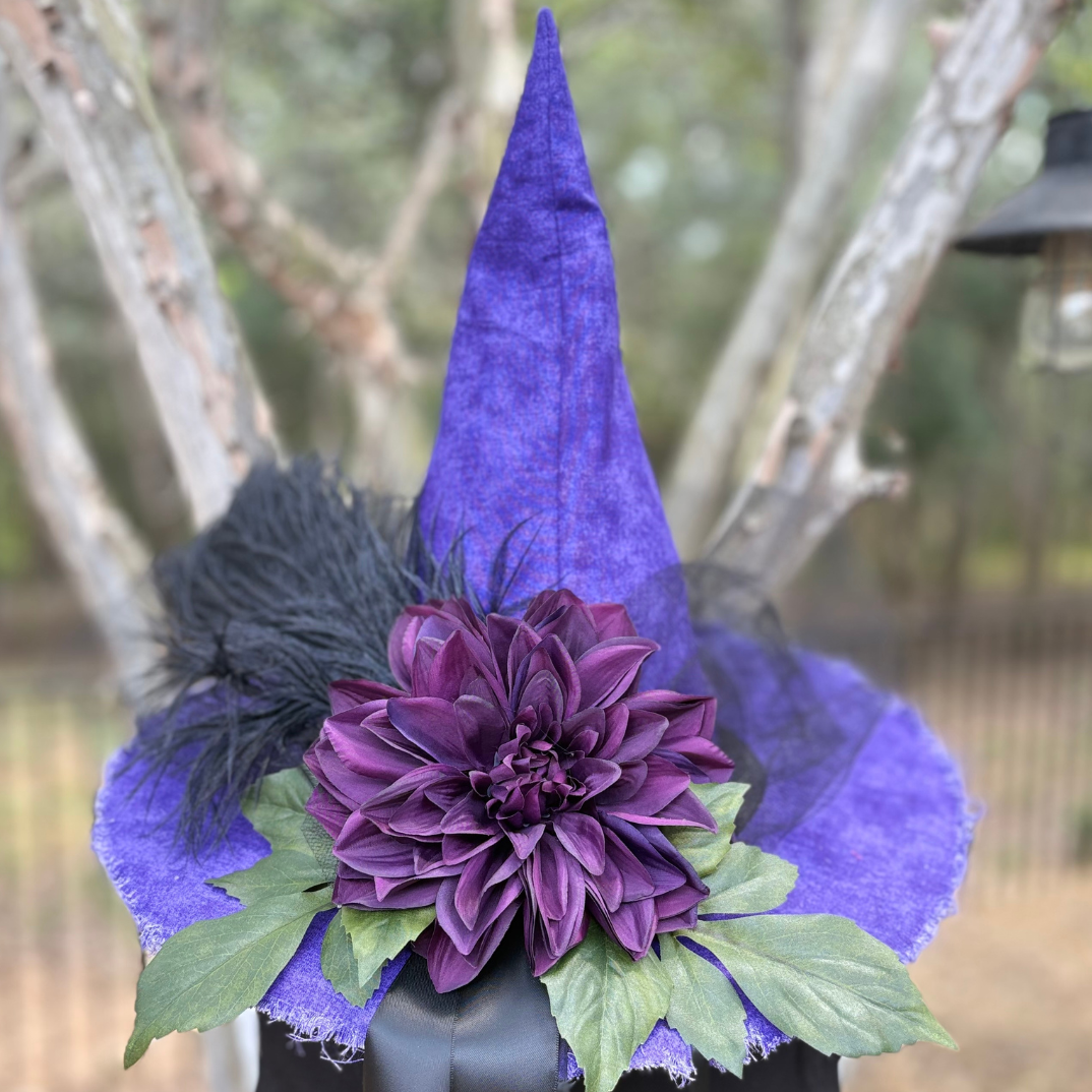 Purple fabric witch hat decoration with a large purple flower, green leaves, and black feathers, displayed outdoors on a black container.